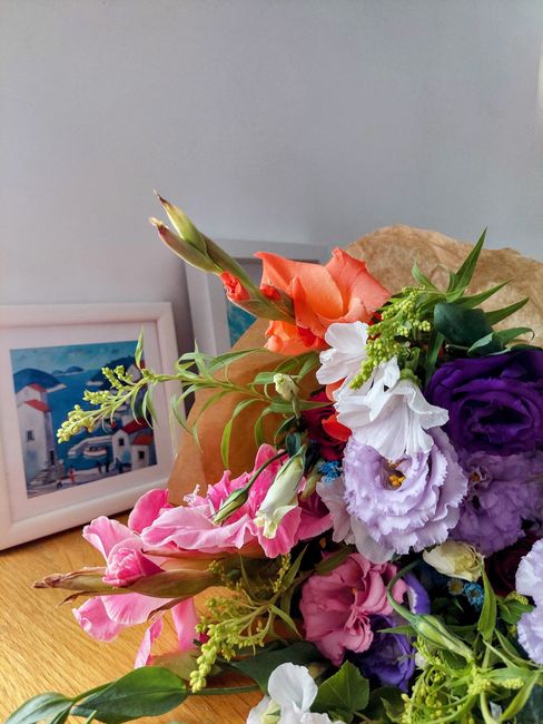 A vibrant bouquet of fresh flowers arranged with peach, white, pink, purple, and green blooms including lisianthus, delphiniums, and other mixed floral varieties, wrapped in brown kraft paper and plac