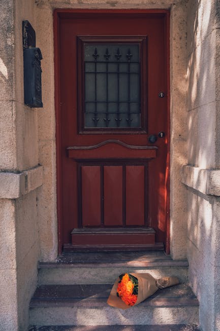 A close-up view of a red wooden front door with a small, decorative iron grille window. In front of the door, on the stone step, lies a bouquet of fresh flowers wrapped in brown paper. The bouquet fea