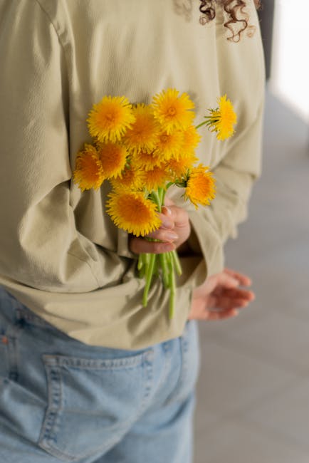 A person wearing a beige long-sleeve top and light blue jeans is holding a bouquet of vibrant yellow chrysanthemums with lush, green stems. The flowers are fresh, with full, textured petals, and are a