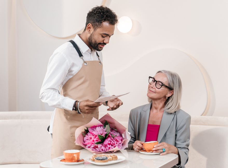 A florist from floristmortlake.co.uk presenting a bright bouquet of fresh pink peonies and chrysanthemums wrapped in pink tissue paper to an elderly woman with silver hair, black glasses, and a light
