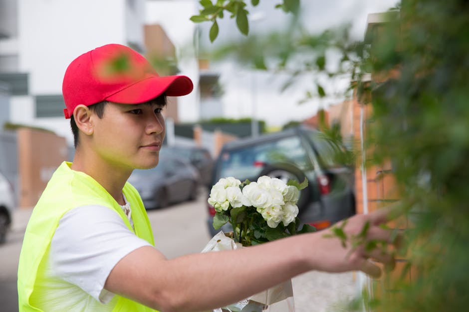 A flower delivery person wearing a red cap and a neon yellow vest is placing a bouquet of fresh white roses and hydrangeas against a brick wall. The bouquet is wrapped in light-colored paper, and the