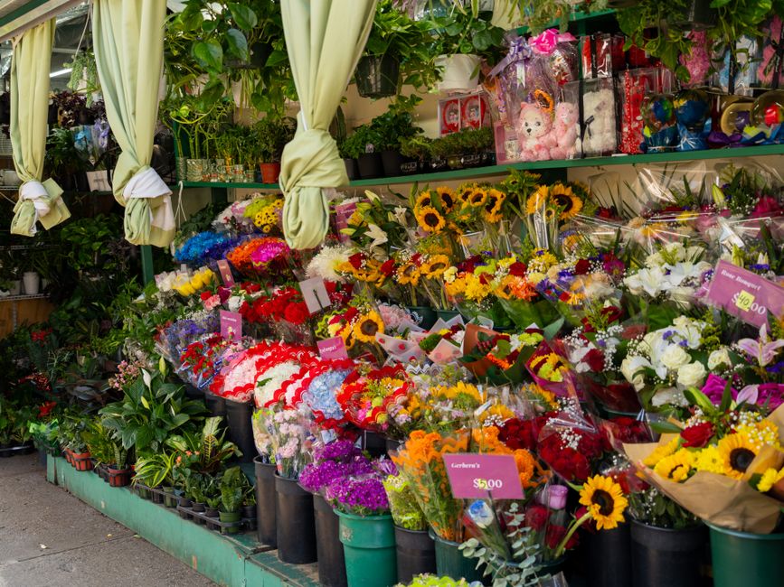 A vibrant flower stall displaying a variety of fresh flowers arranged in black buckets and wrapped bouquets. Bright yellow sunflowers, white lilies, and colorful mixed blooms are visible, with some wr